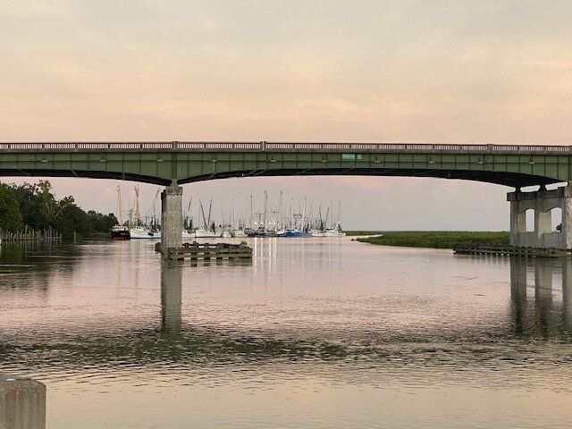 Bridge over calm river at sunset, with sailboats in the distance.