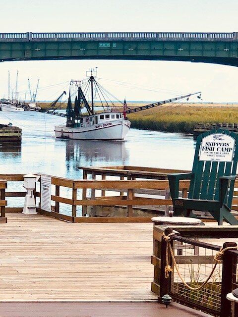 Fishing boat in a canal under a bridge, viewed from a wooden dock with railings and chairs.