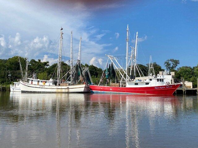 Two sailboats docked on a calm river, one white and one red, with trees and blue sky reflected in the water