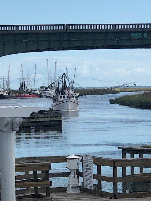 Fishing boat in a harbor under a bridge, with cranes and docked boats in the background.