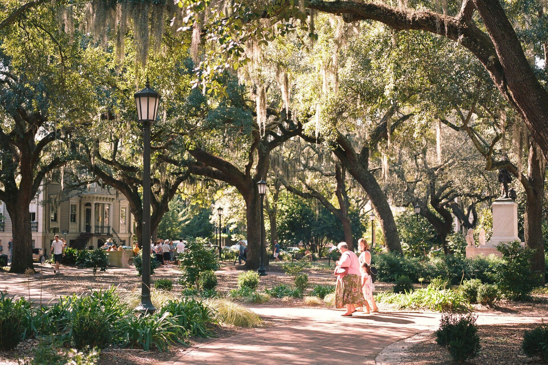 Shaded park path with large trees, a lamppost, and two people walking in pink dresses