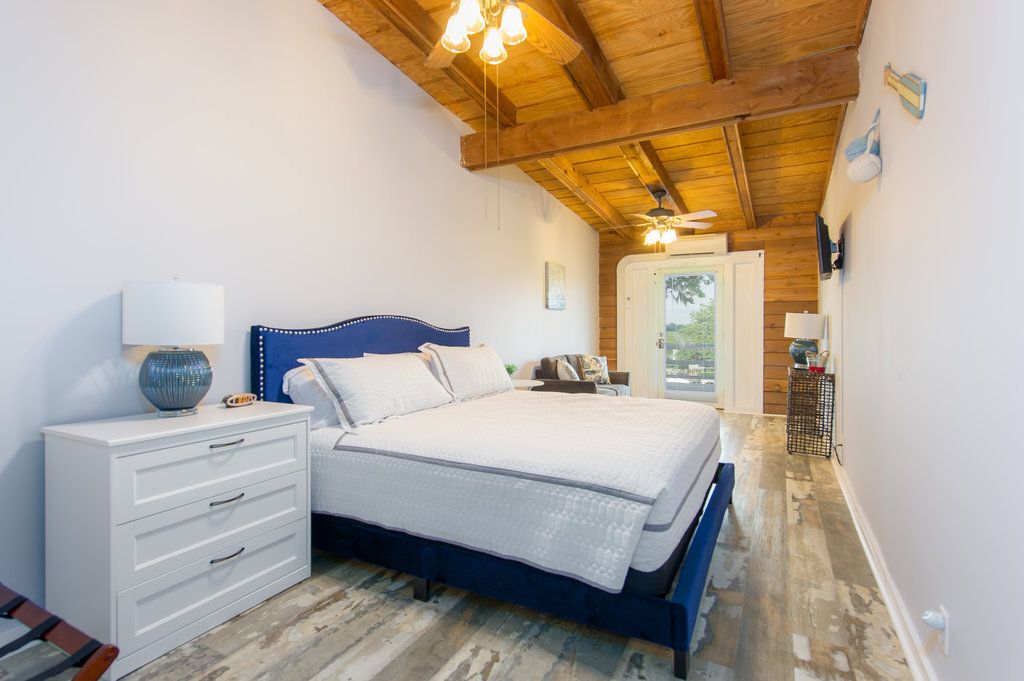 Bright bedroom with blue bed, white dresser, wood-beamed ceiling, and large window.