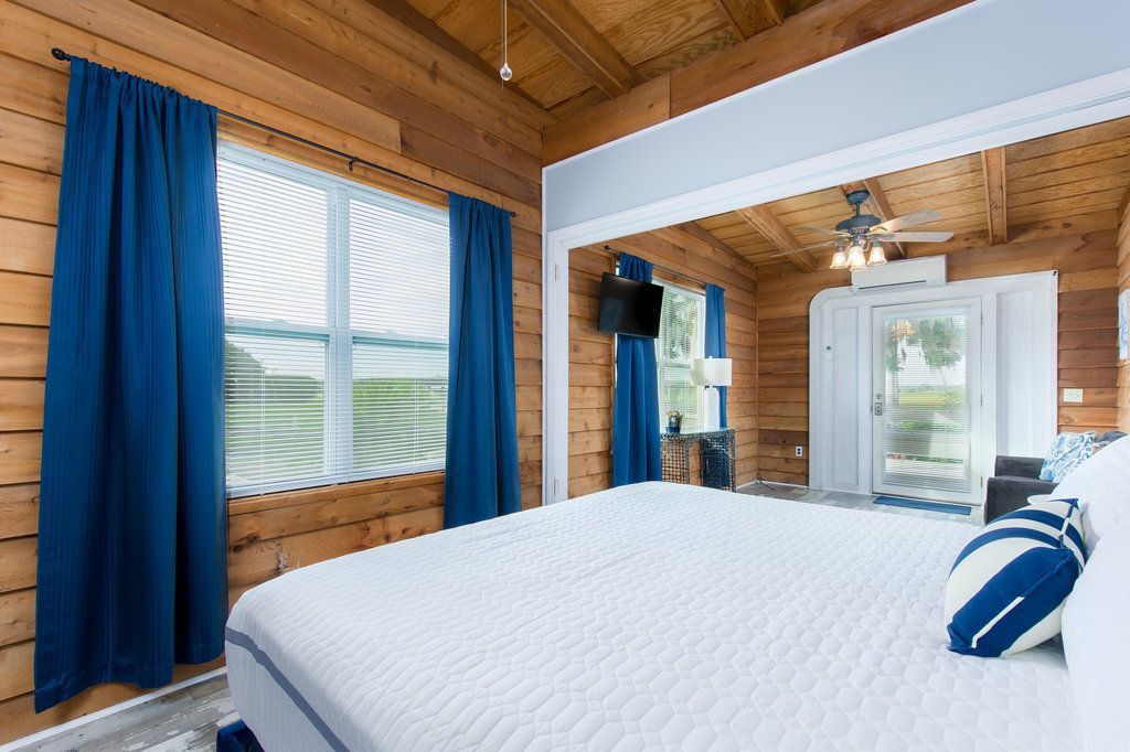 Bright bedroom with wooden ceiling, white bed, blue curtains, and a large mirror near a window.