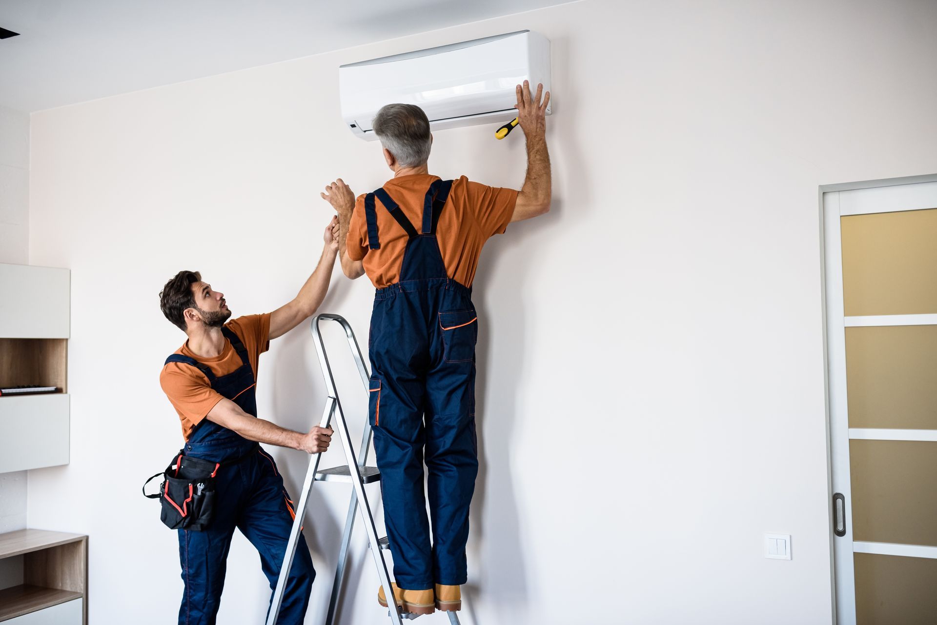 Two technicians in dark overalls, using a ladder, are performing an AC installation.