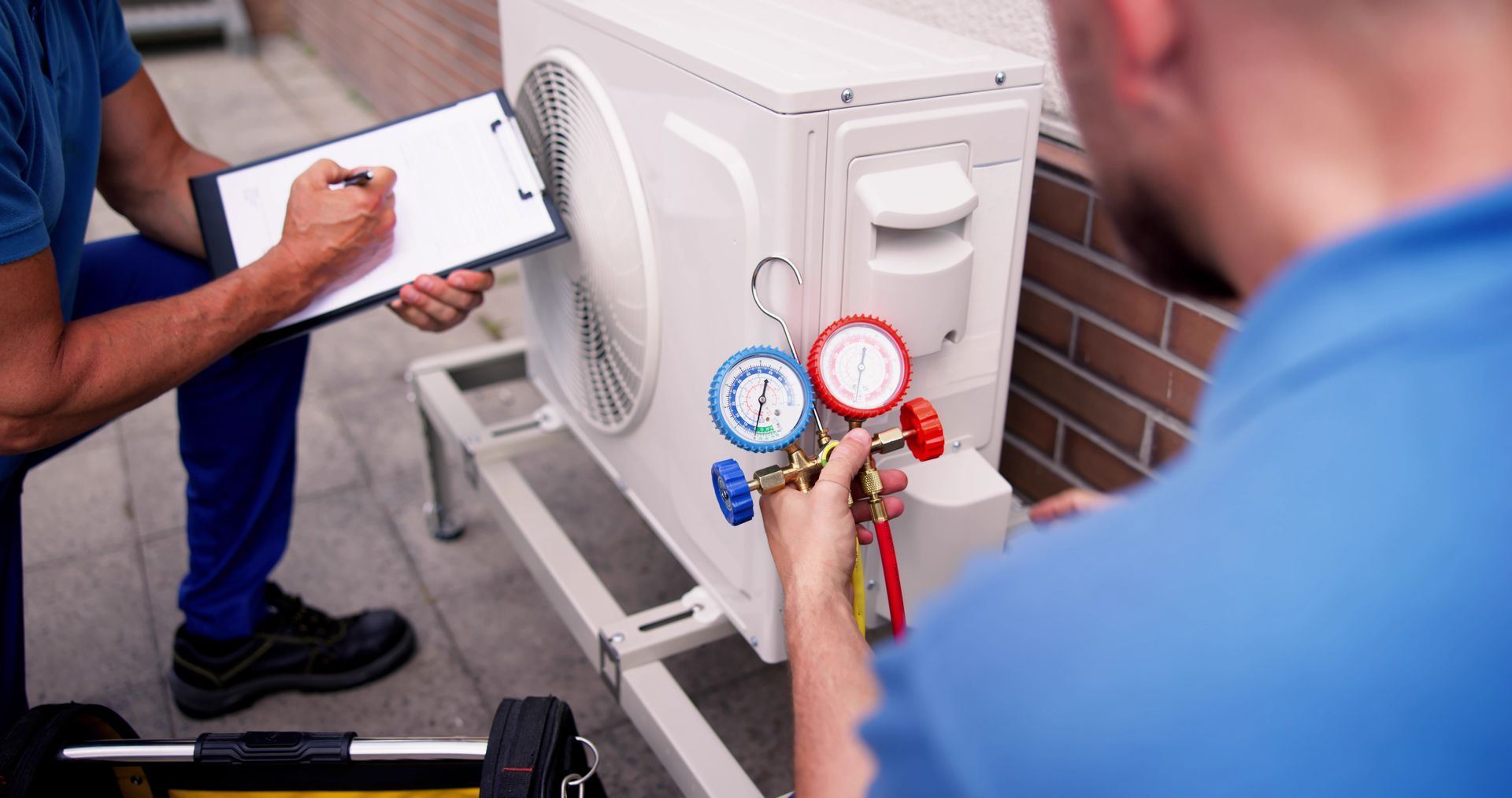 Two AC workers checking an AC unit outside a building, taking notes.