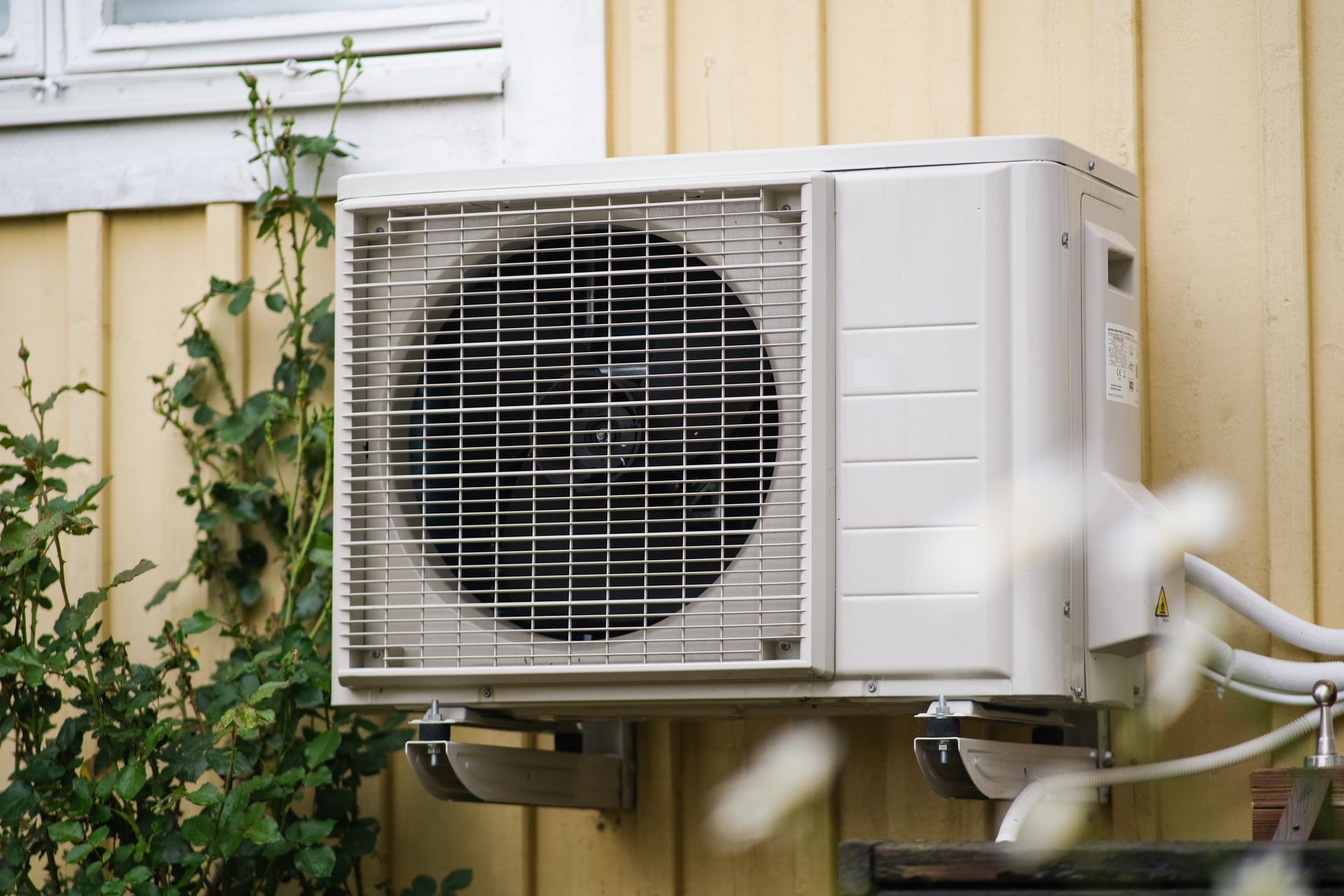 An outdoor beige AC unit mounted on a yellow wall near plants.