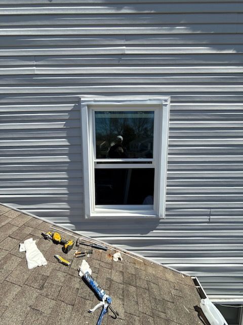 Window in gray siding above shingled roof, with roofing tools scattered nearby.