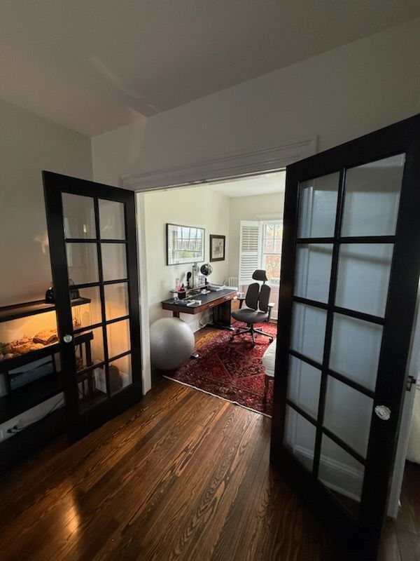 French doors open to a cozy home office with a desk, red rug, and wooden floor.