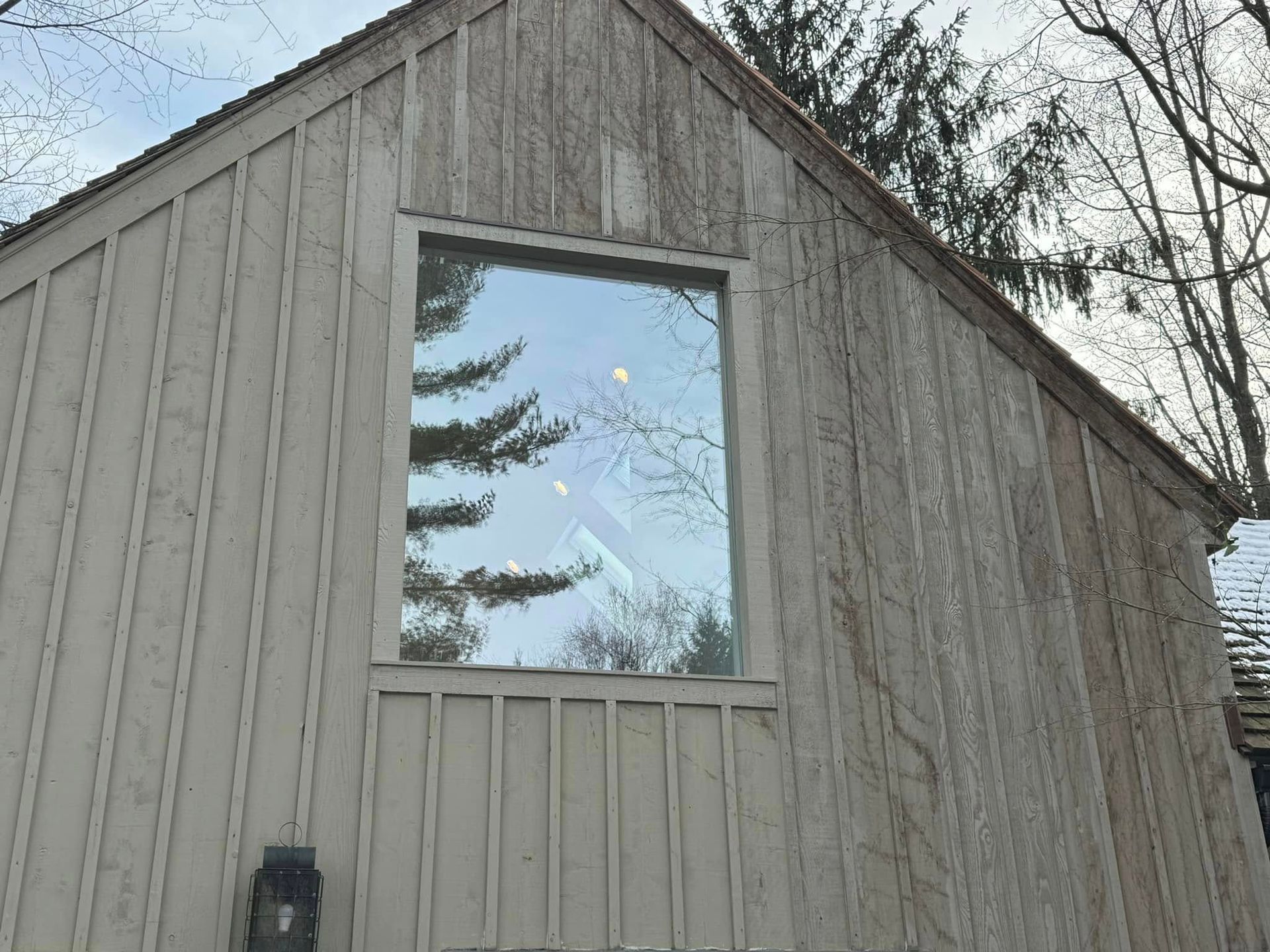 A light-colored wooden building with a large window reflecting trees and sky.