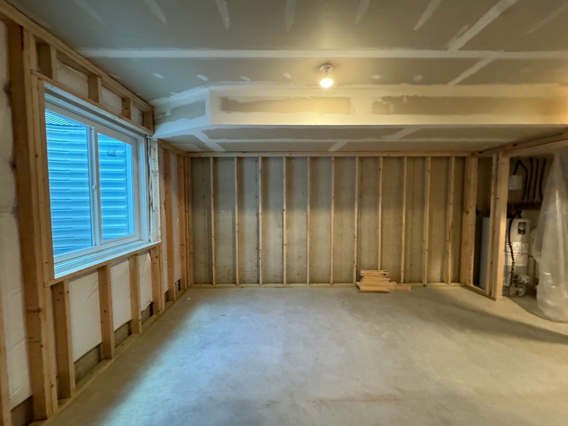 Framed basement room with exposed wooden studs, drywall partially installed. Window on left.