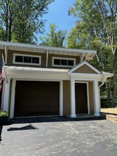 Detached two-car garage with white trim, dark doors, and a paved driveway under trees