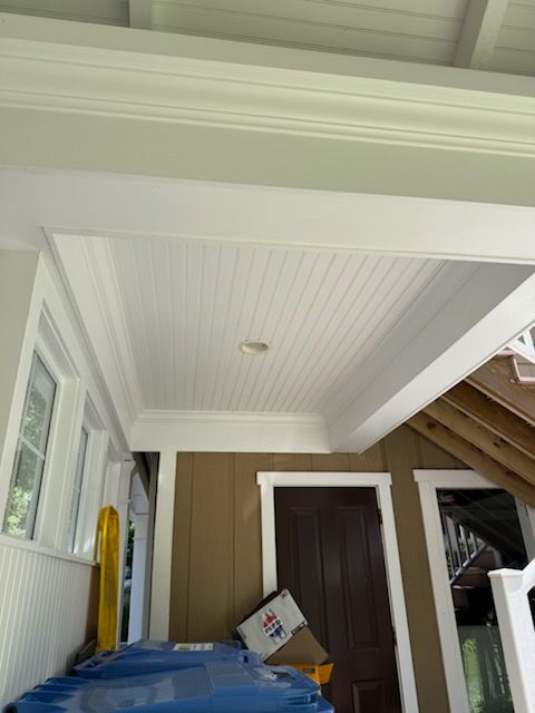 Covered porch ceiling with white beadboard trim, brown door, windows, and a tarp-covered table.