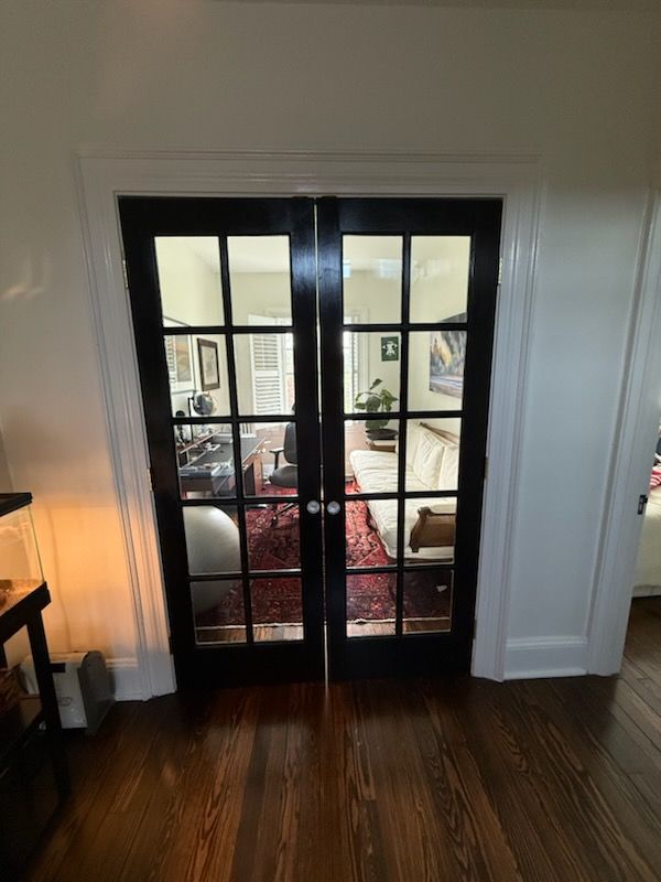 Black French doors with glass panes open to a cozy room with a red rug and lamp.