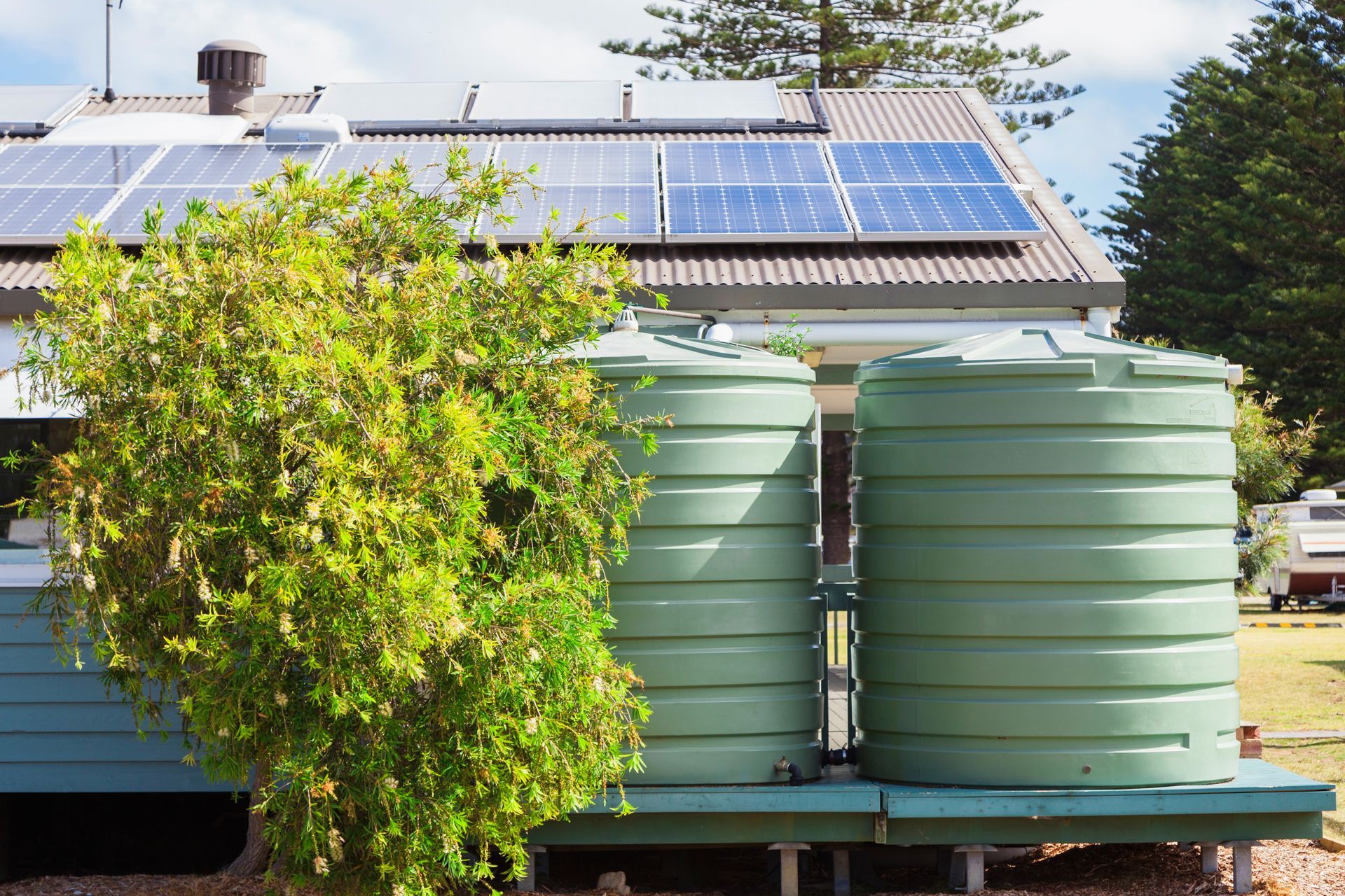 Solar panels on a roof behind green rainwater tanks and a leafy bush outside a house