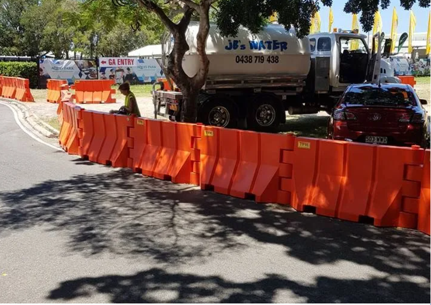 Orange barricades line a street beside a parked water truck and red car under trees.