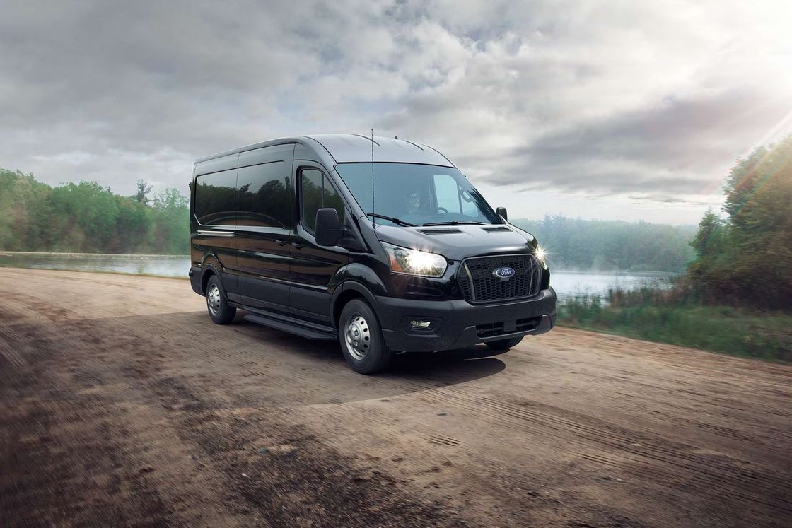 Black Ford Transit van parked on a dirt road next to a body of water. Overcast sky.