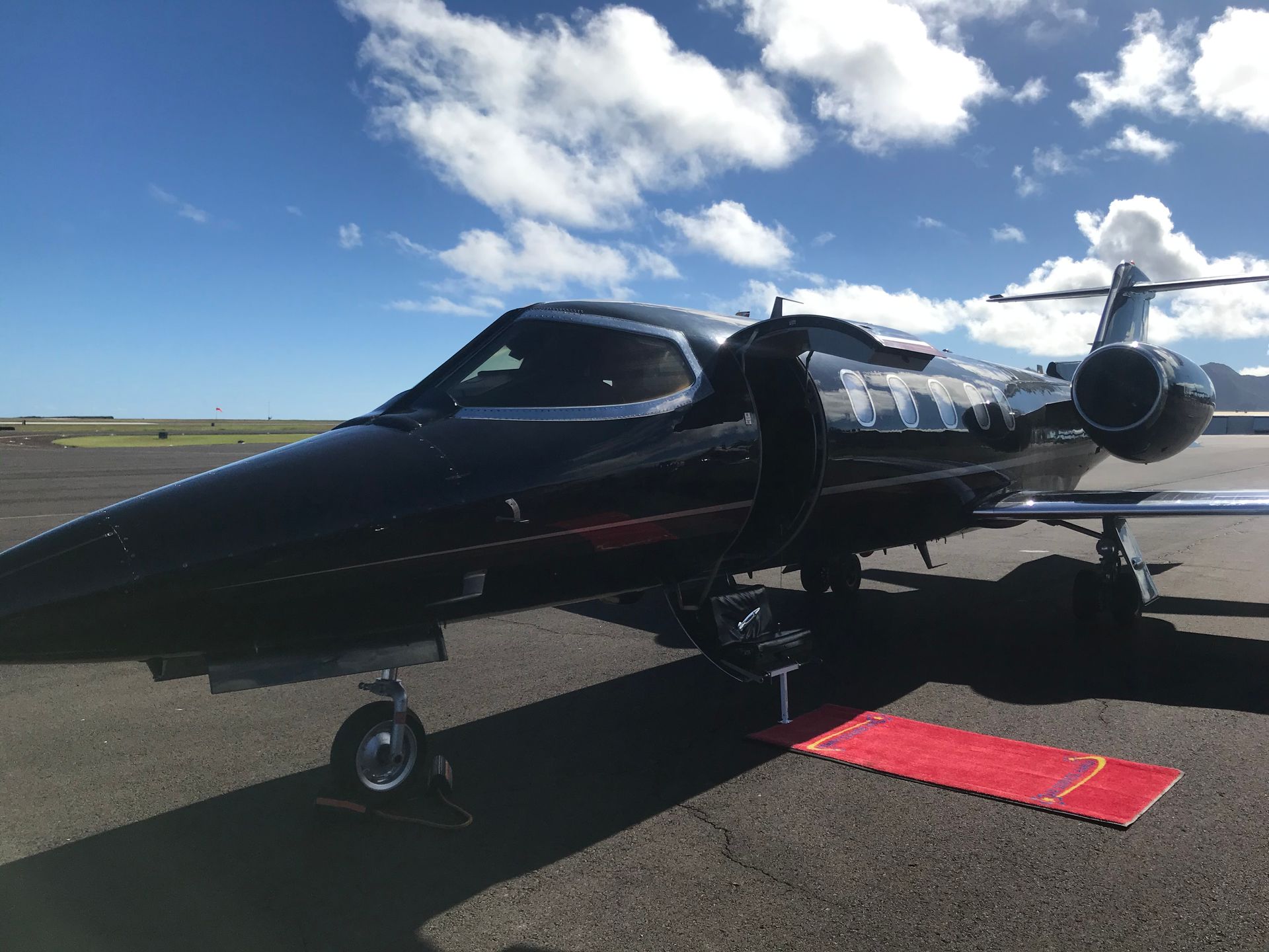 Black private jet on the tarmac with red boarding step; blue sky with clouds in background.