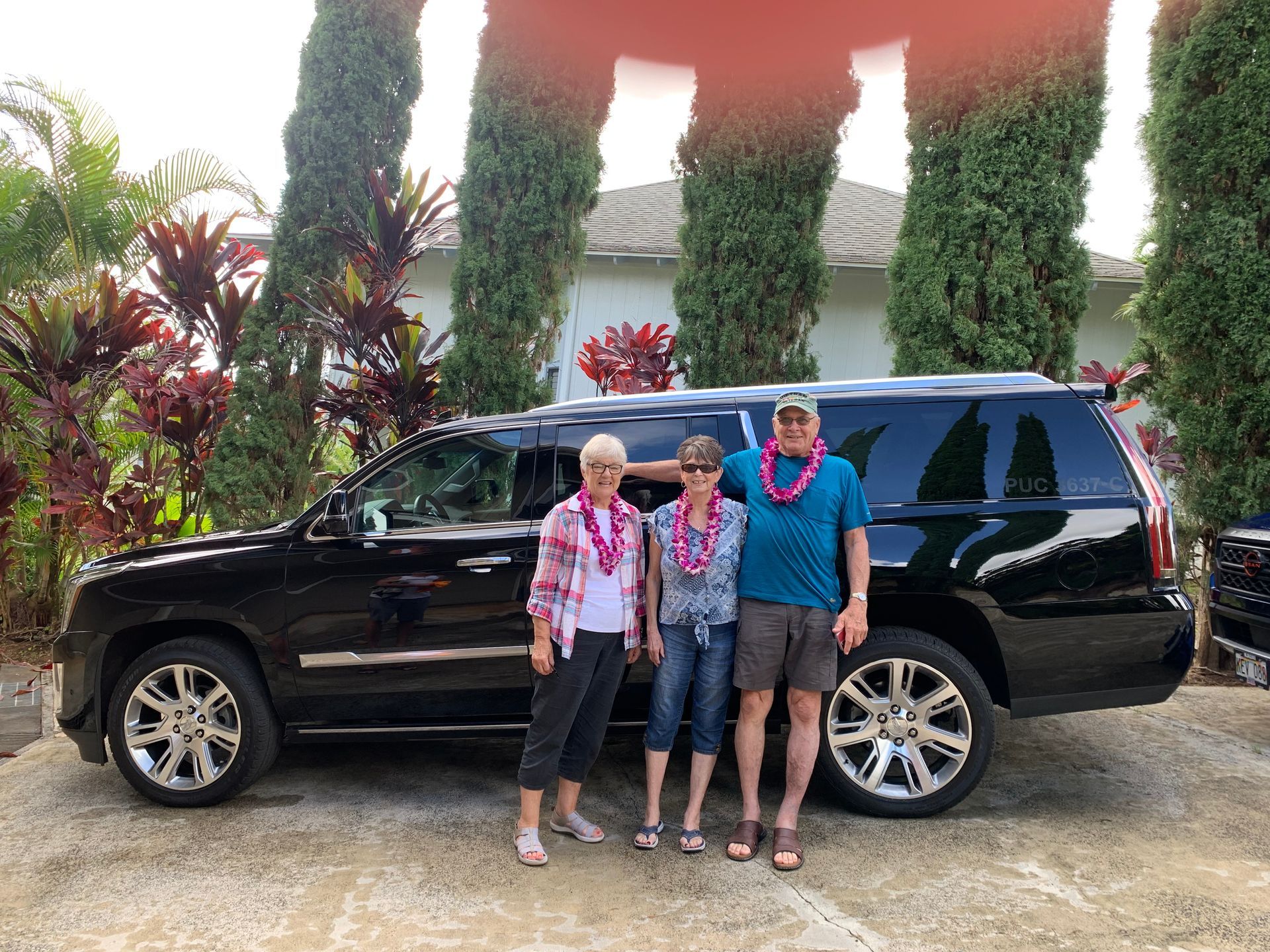 Three people in leis pose next to a black SUV in front of a house.
