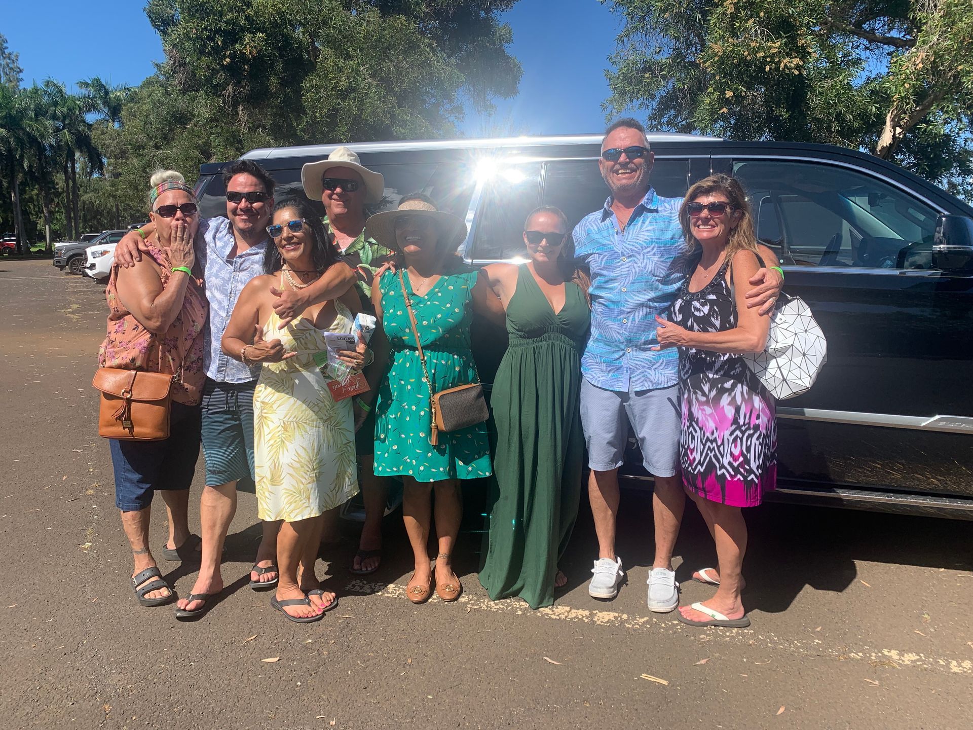 Group of nine people posing in front of a black vehicle, smiling under sunny skies.