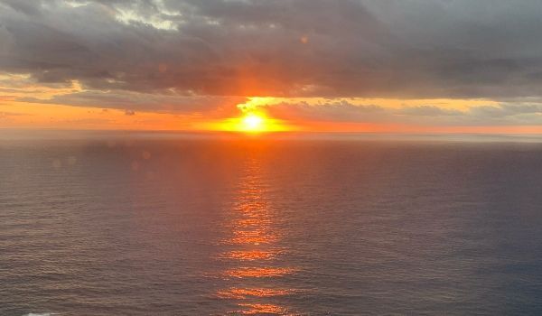 Sunset over ocean; golden sun reflects on water, obscured by clouds.