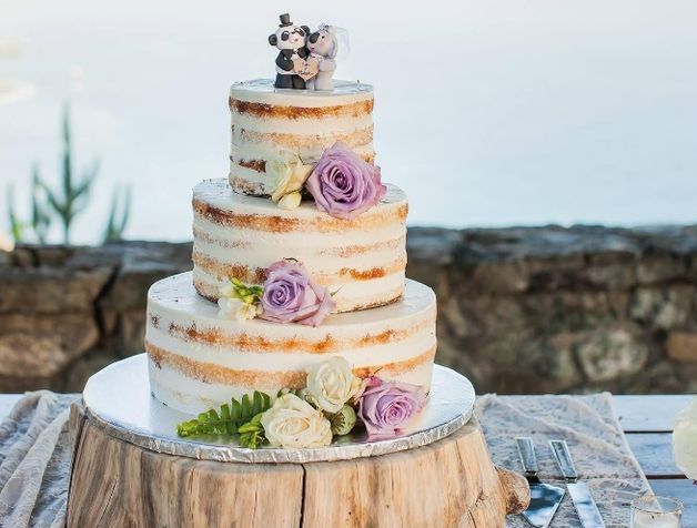Three-tiered naked wedding cake decorated with purple roses and panda toppers on a rustic wood stand.