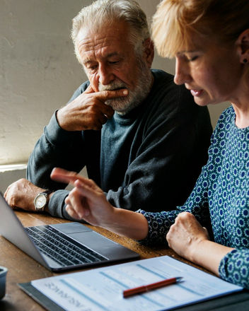 Two people discuss a laptop at a table, with notes and a pen in the foreground.