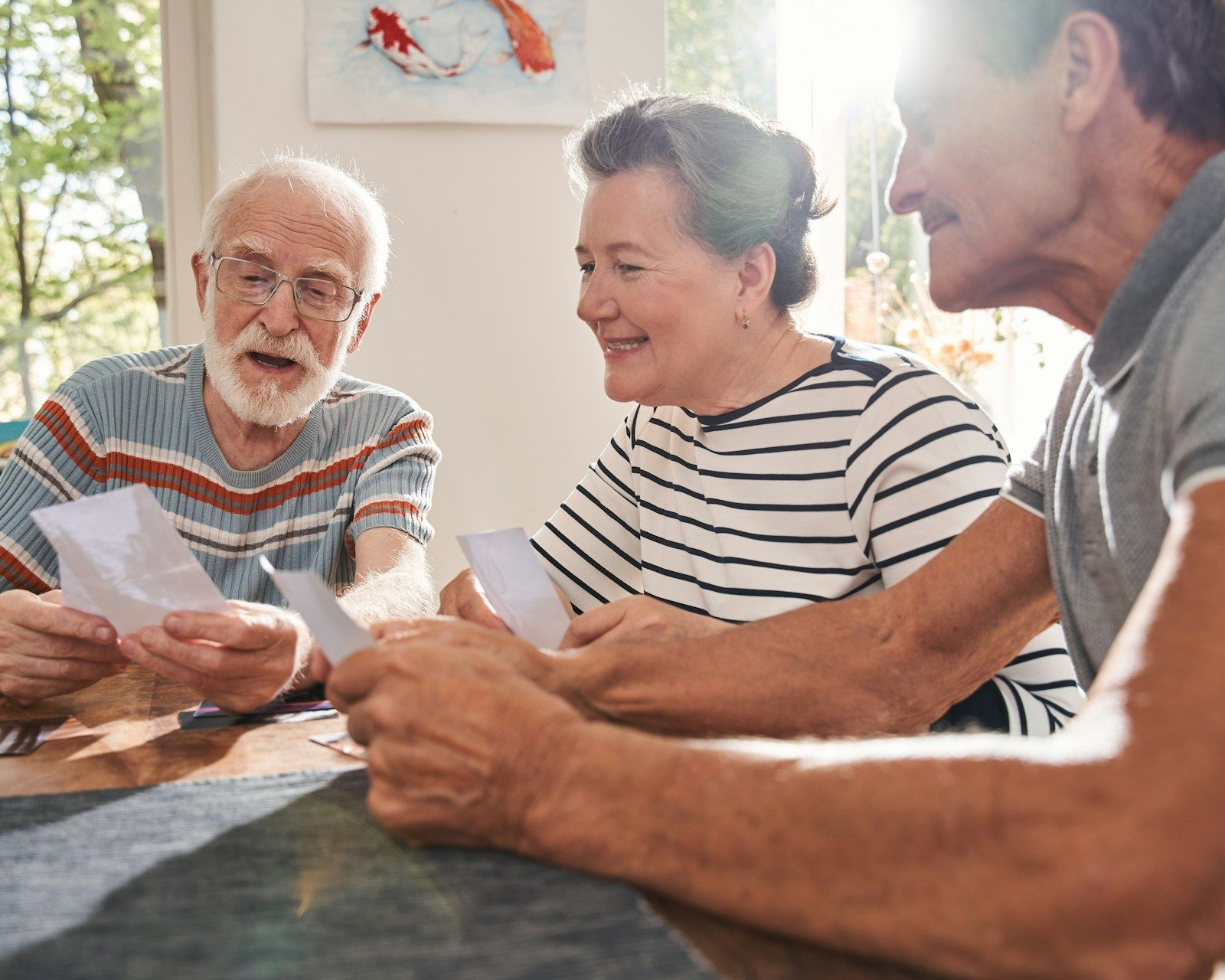 Three people talking and holding papers at a sunlit table indoors.