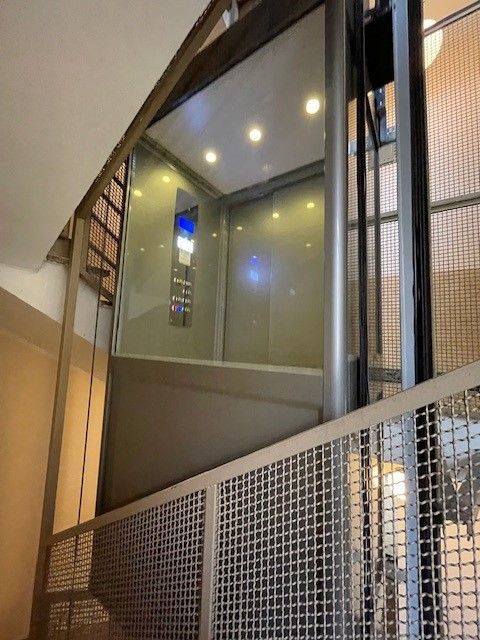 Ascensore con pareti in vetro, all'interno di un vano metallico. Pannello di controllo visibile. Ben illuminato con luci sospese.