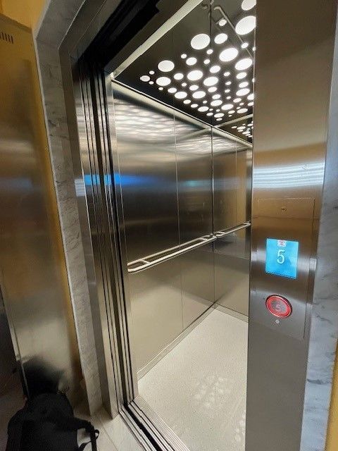 Ascensore con porte aperte, interni in acciaio inox. Pulsante per il 5° piano. Soffitto luminoso con lampade circolari.
