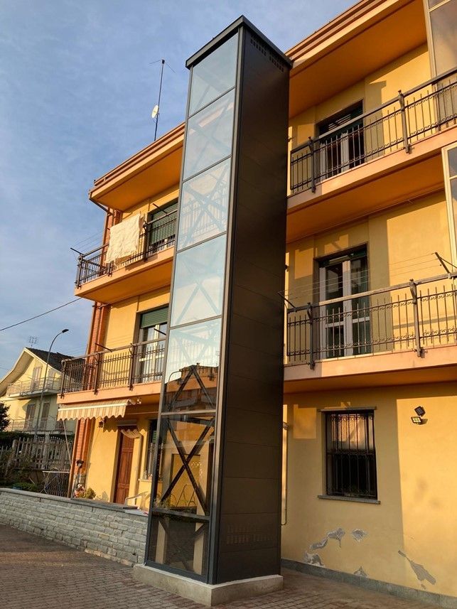 Vano ascensore con pannelli di vetro, montato all'esterno di un edificio giallo. Cielo coperto.