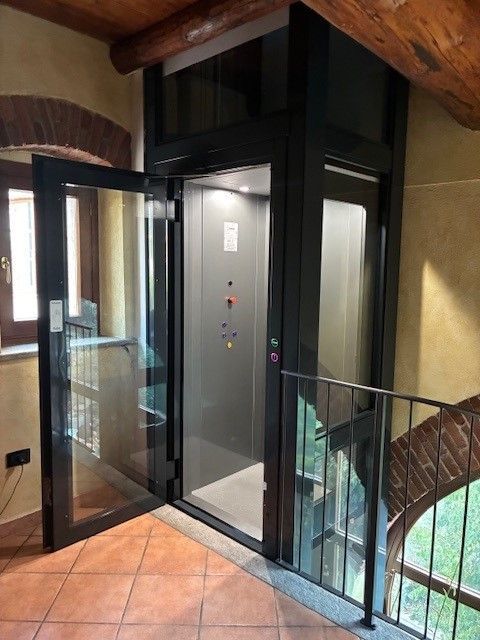 Ascensore con porte in vetro e telaio nero, adiacente a una scala in un edificio con pareti in mattoni.