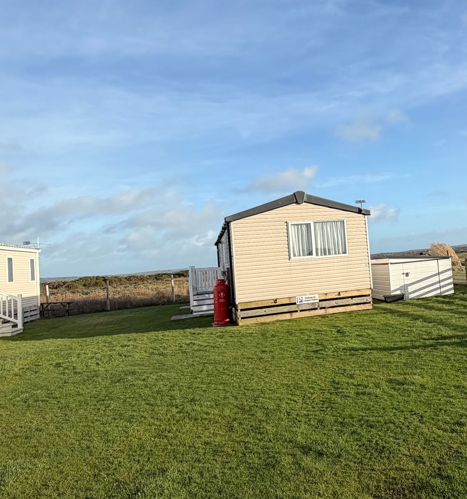 Beige mobile home on a grassy field under a blue sky, with a red post and another trailer nearby.