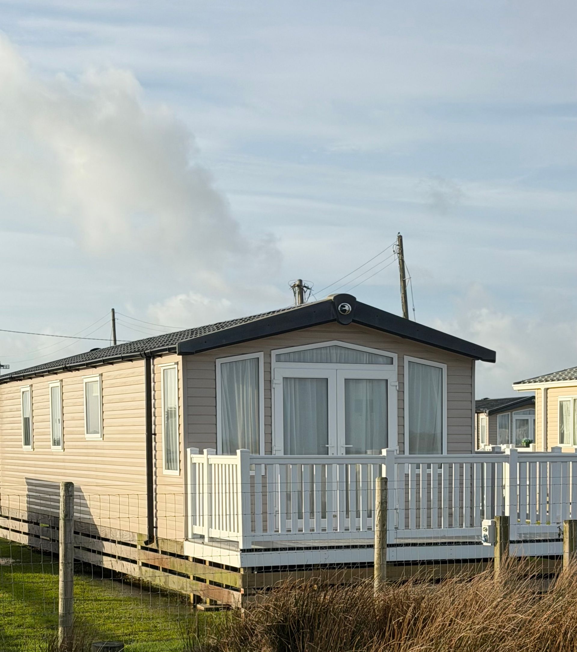 Small beige mobile home with white porch railing under a cloudy sky in a grassy park.