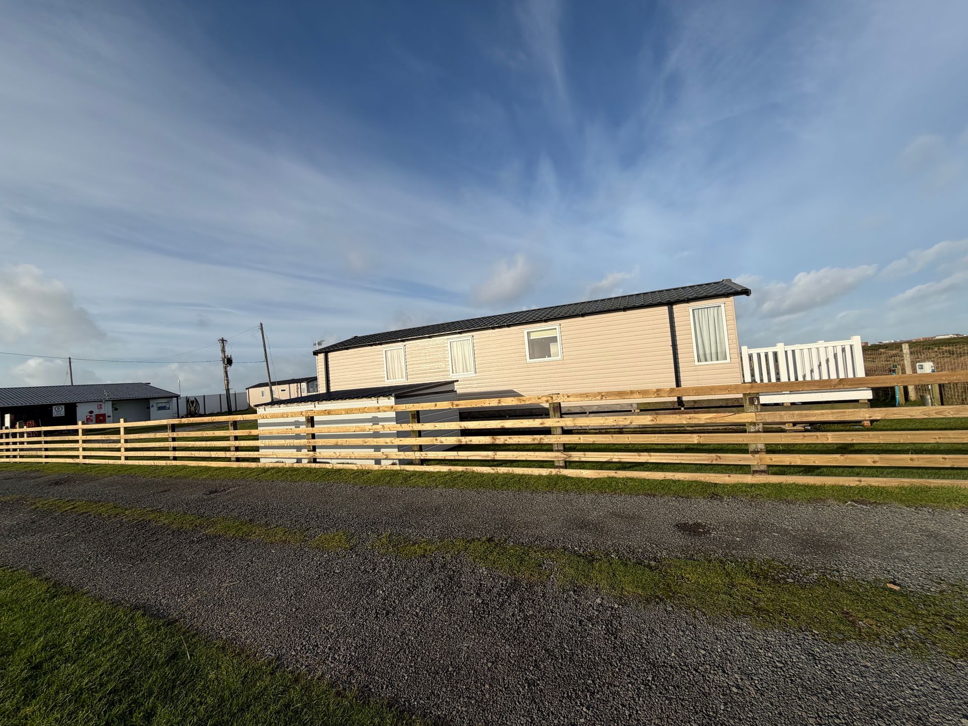 Rural gravel road beside a wooden fence and beige barn under a blue, cloudy sky