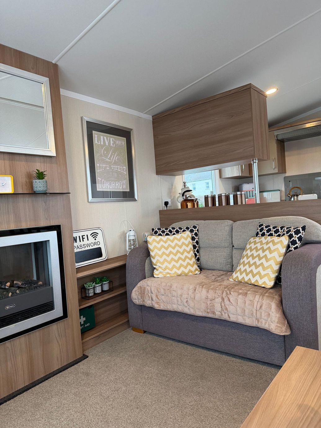Cozy living room with brown sofa, yellow pillows, fireplace, mirror, and kitchen area in the background