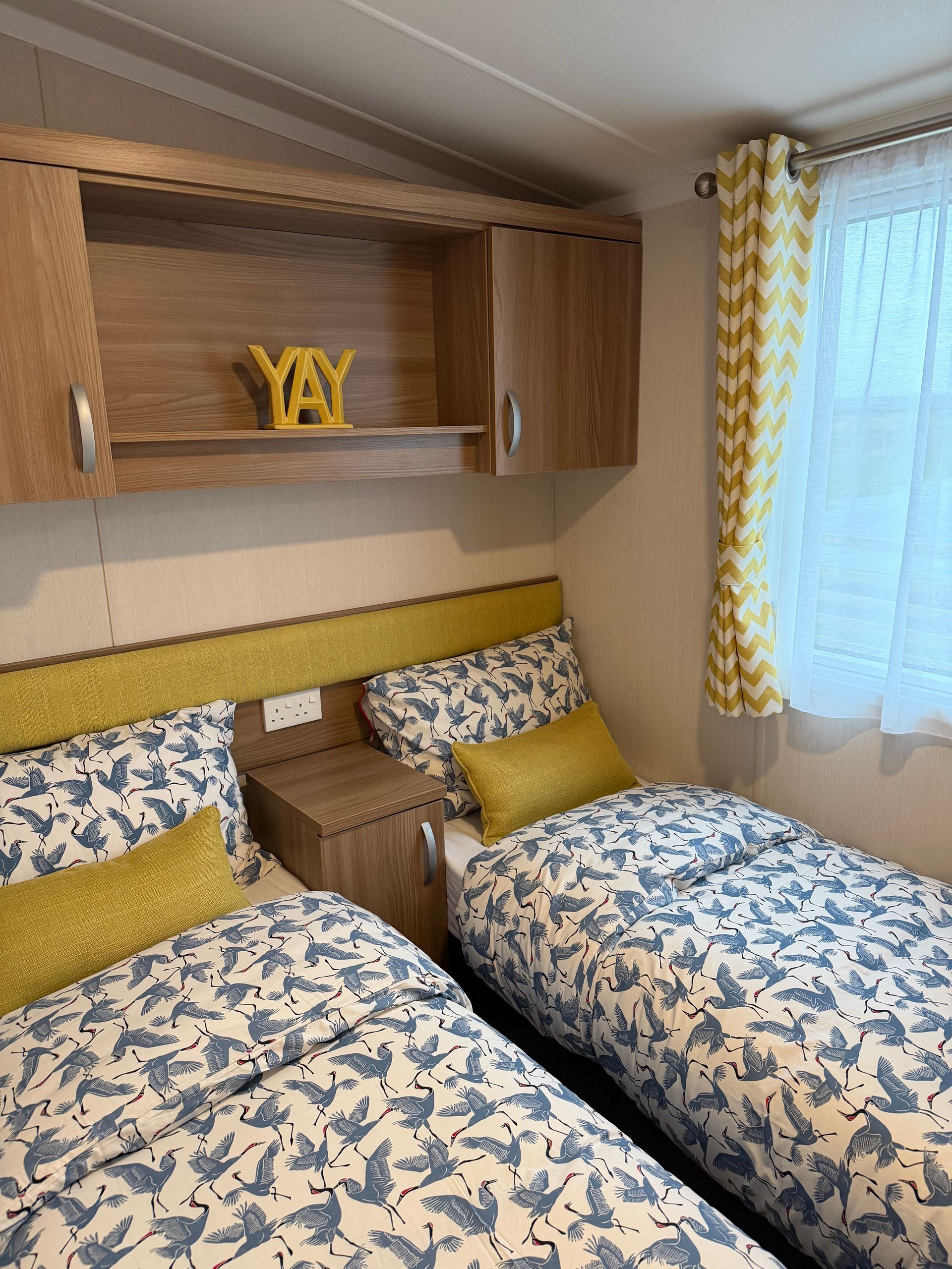Cozy twin bedroom with blue patterned bedding, yellow accents, and a window with sheer curtains.