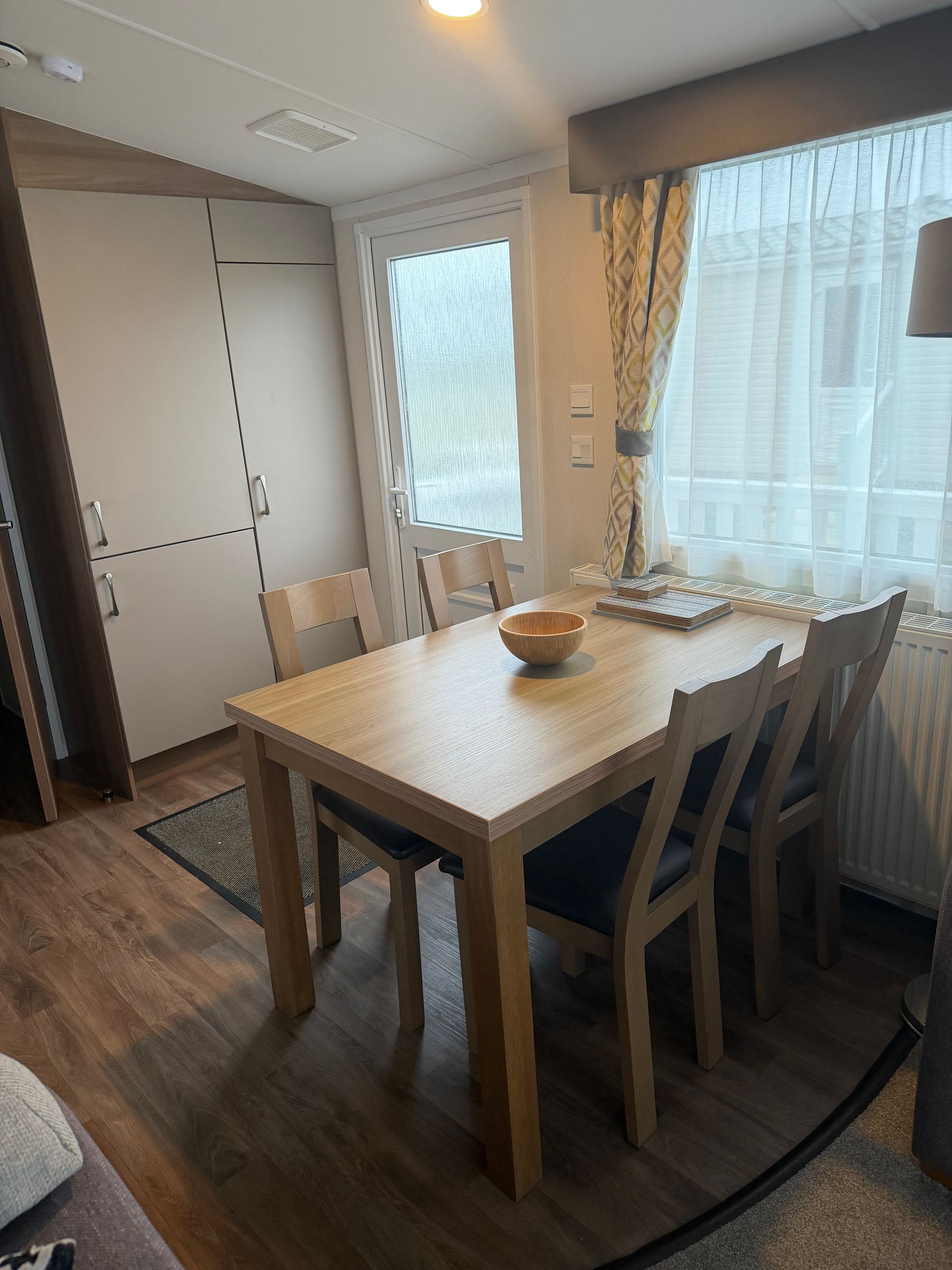 Dining room with wooden table and chairs beside a large window with sheer curtains.