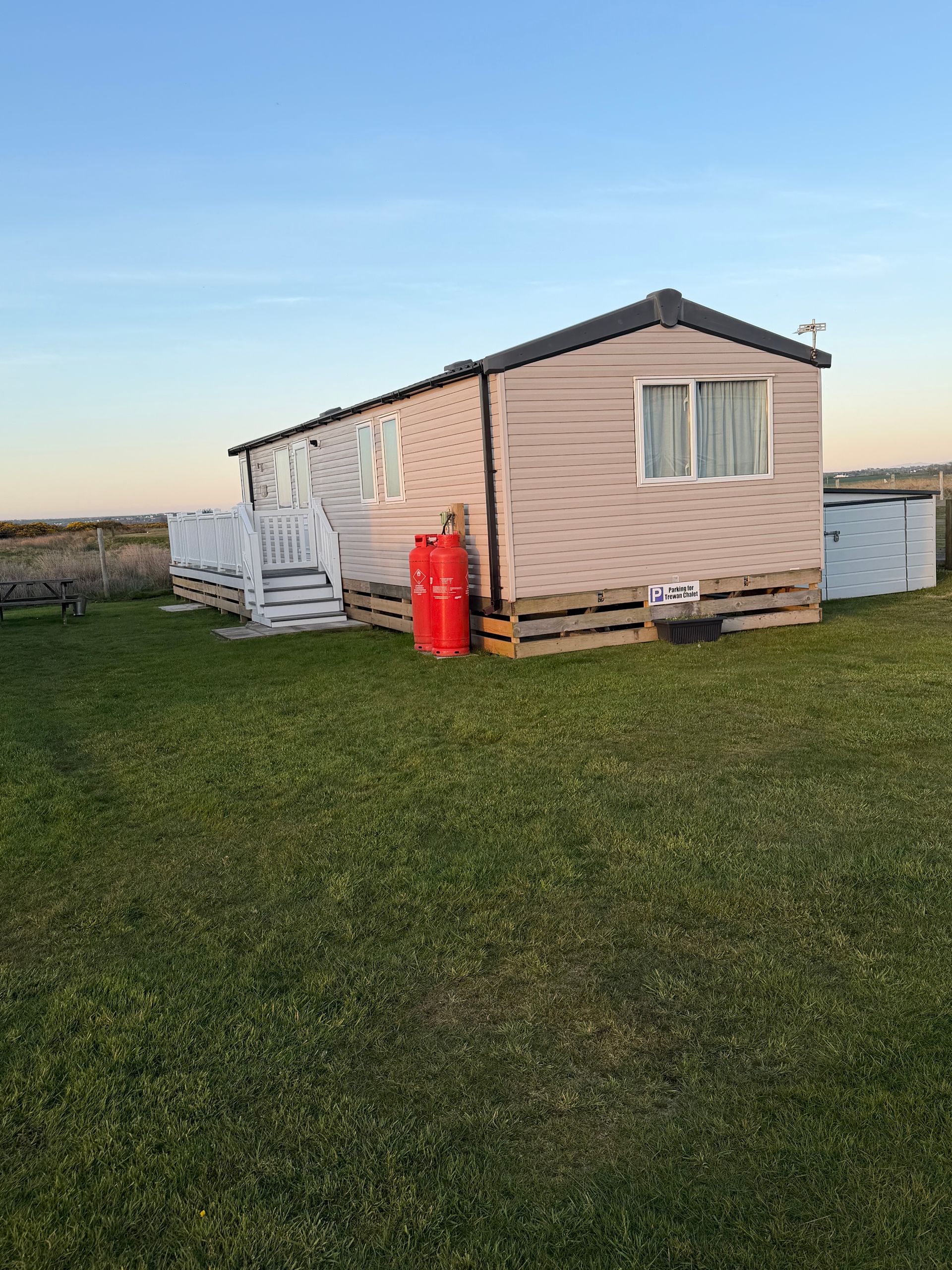 Small beige mobile home on a grassy field at sunset, with white trim and a red gas cylinder beside it