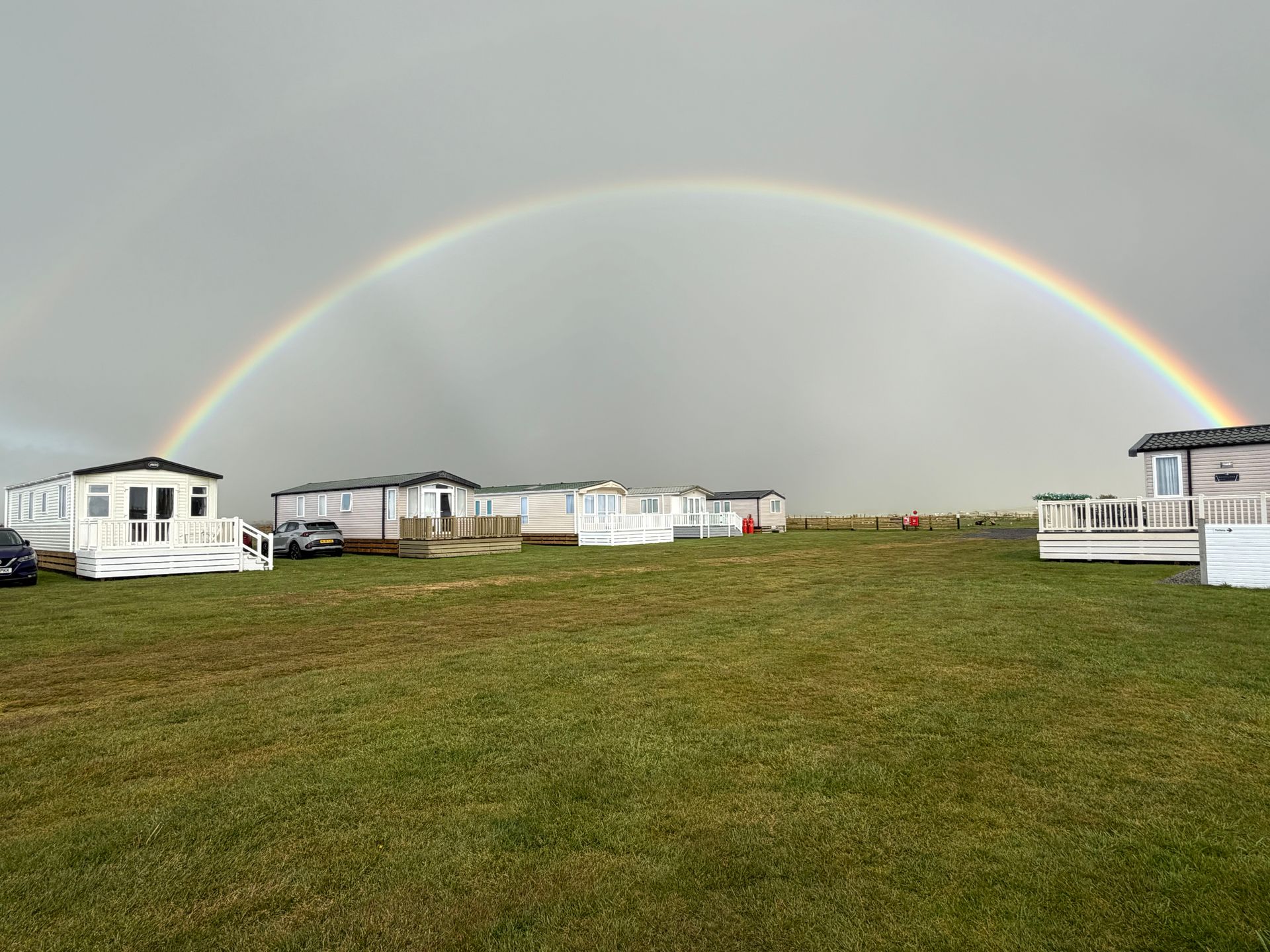Rainbow over a grassy field with white cabins and trailers under a cloudy sky