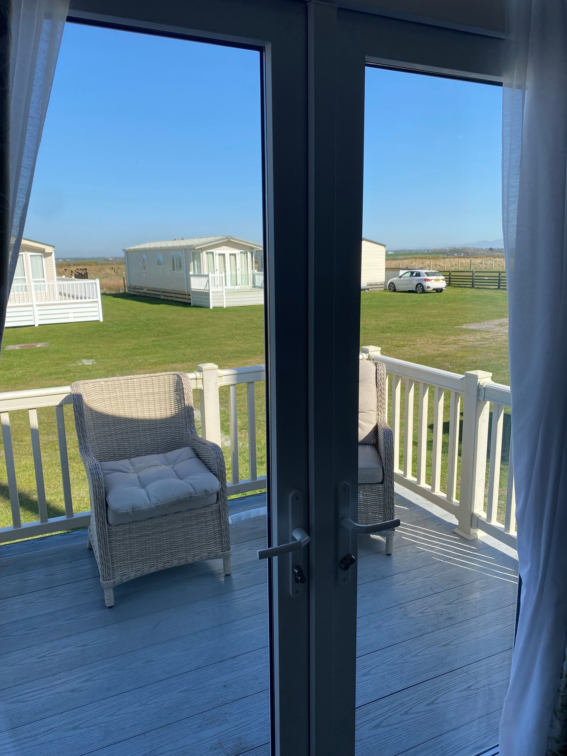View through glass doors to a porch with wicker chair, white railing, grassy field, and parked RVs under a blue sky