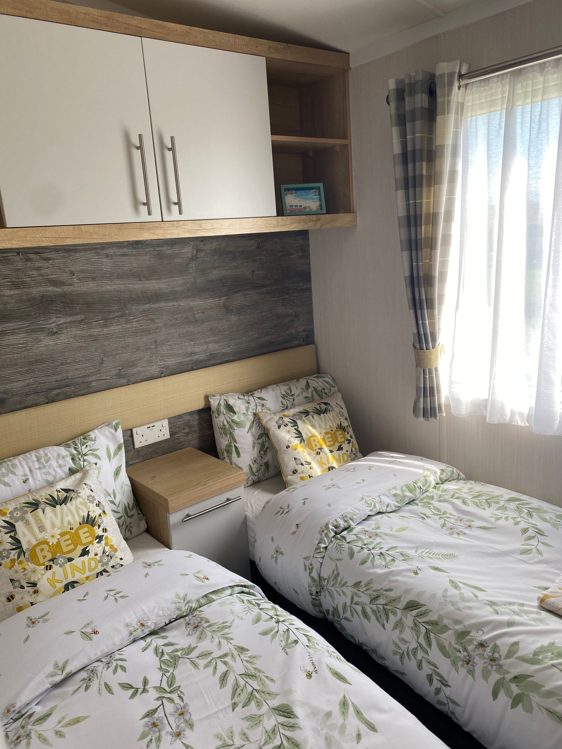 Small bedroom with two twin beds, floral bedding, wall cabinets, and a bright window with curtains.