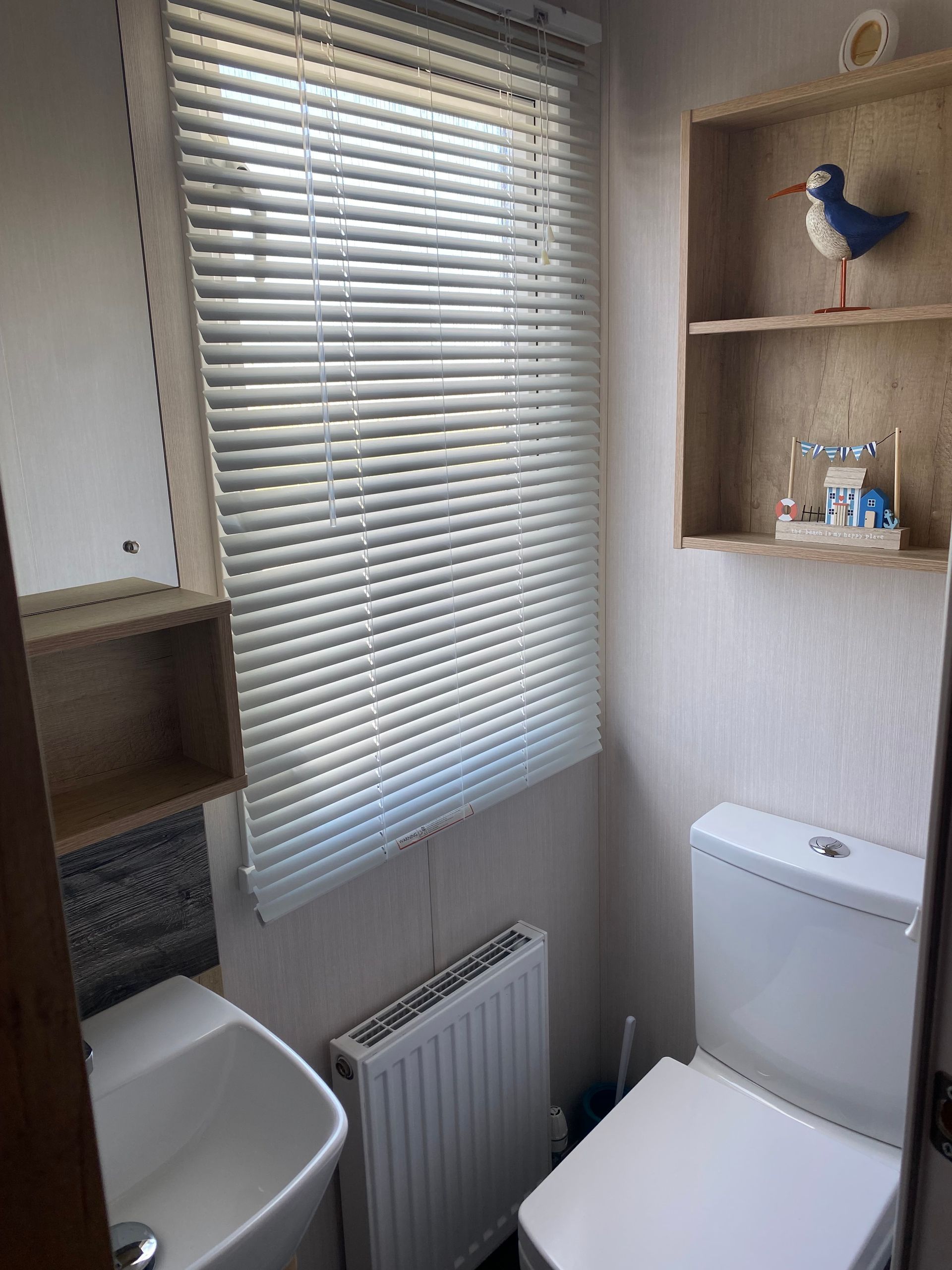 Small bathroom with toilet, sink, radiator, window blinds, and wooden wall shelves.