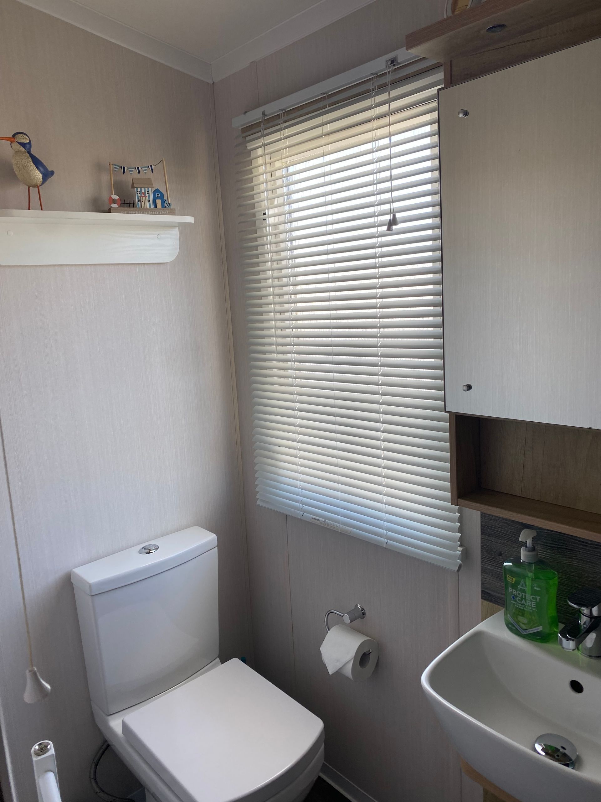 Small bathroom with a toilet, sink, window blinds, and a wall shelf with toiletries.