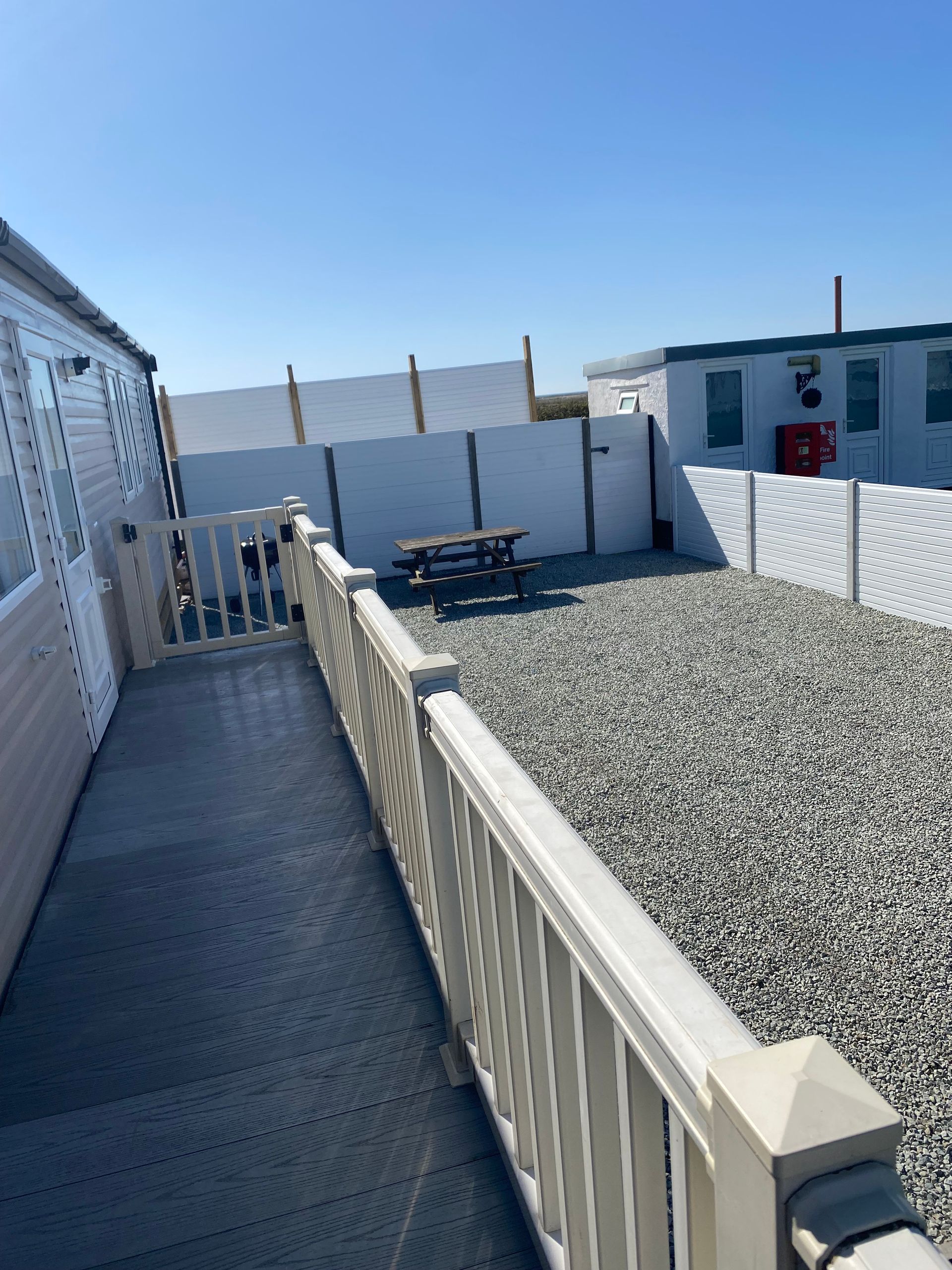 Narrow deck walkway beside a white building, with gravel yard and picnic table in bright sunlight.