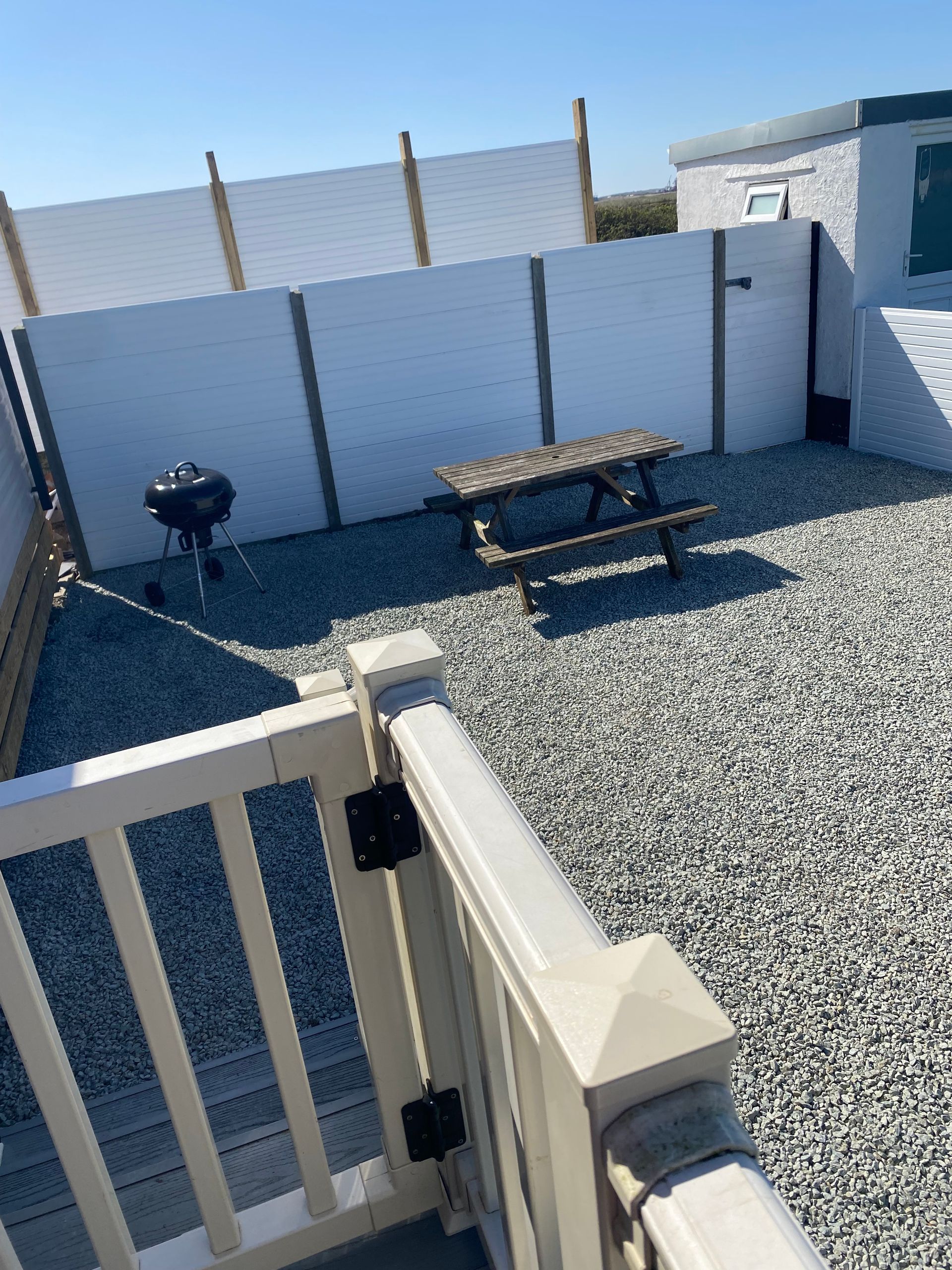Small gravel patio with a picnic table, grill, white fence, and a gate in the foreground.
