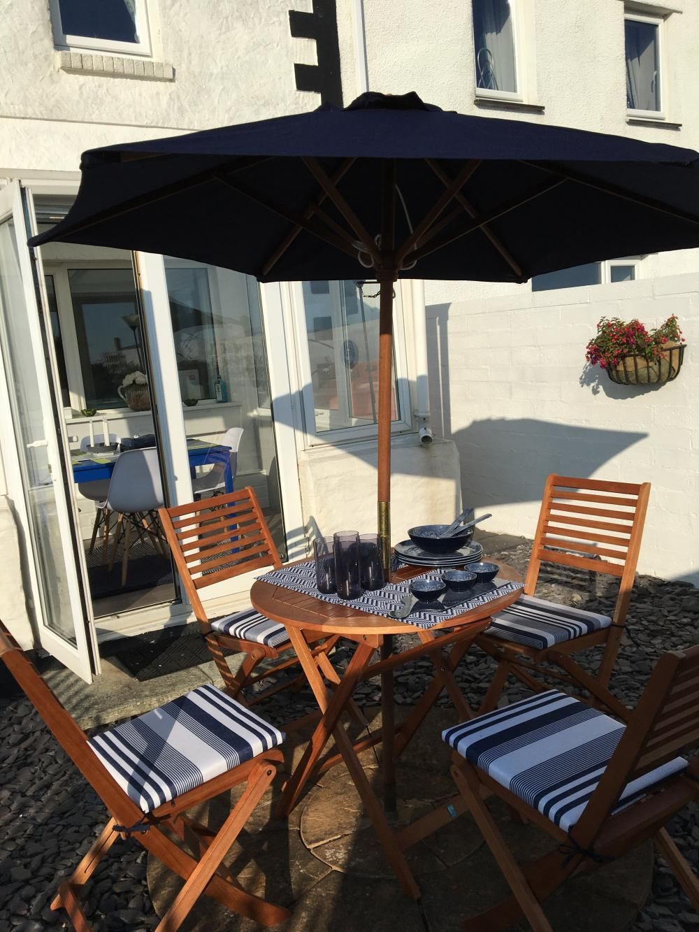 Outdoor patio table with four wooden chairs, a black umbrella, and a tea set beside a house
