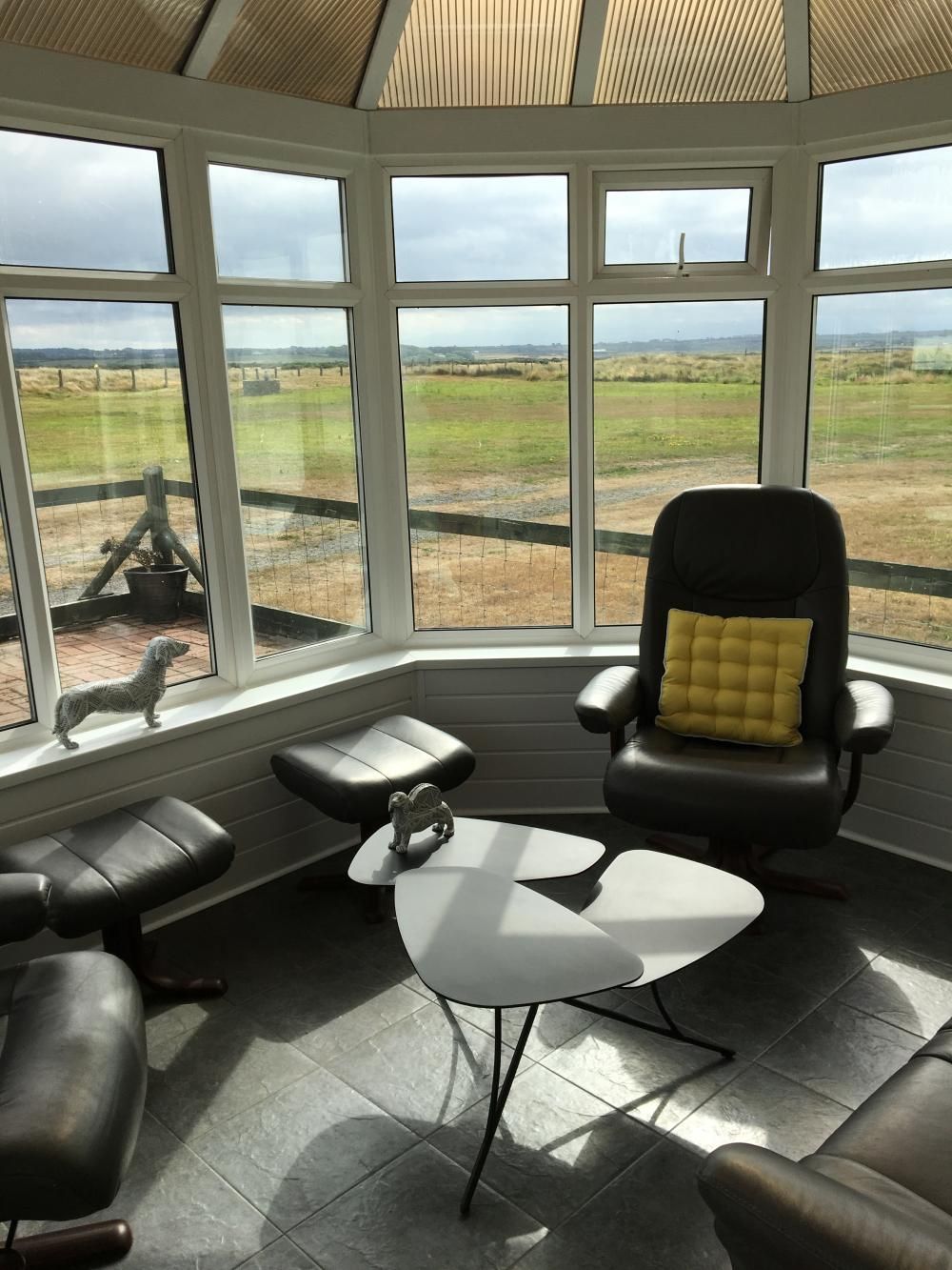 Bright sunroom with large windows, leather chairs, and a small white table overlooking a grassy field.