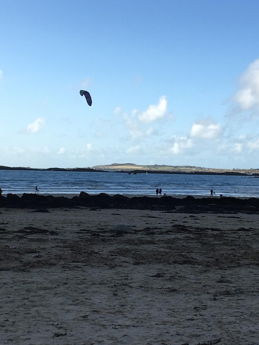 Beach with dark sand, blue water, distant island, and a kite or parasail in a partly cloudy sky
