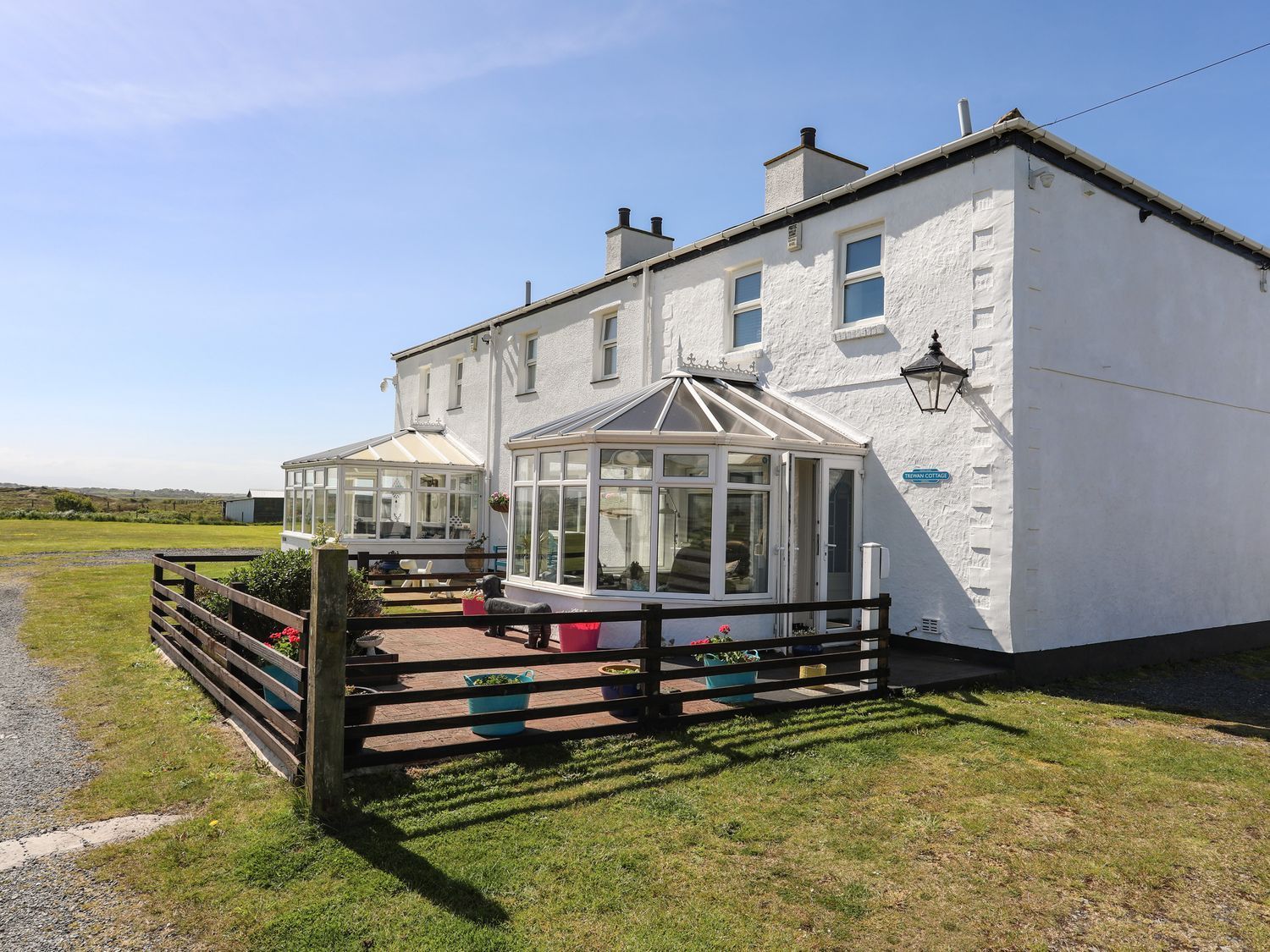 White coastal cottage with sunroom, black fence, and grassy yard beside a gravel lane under a blue sky