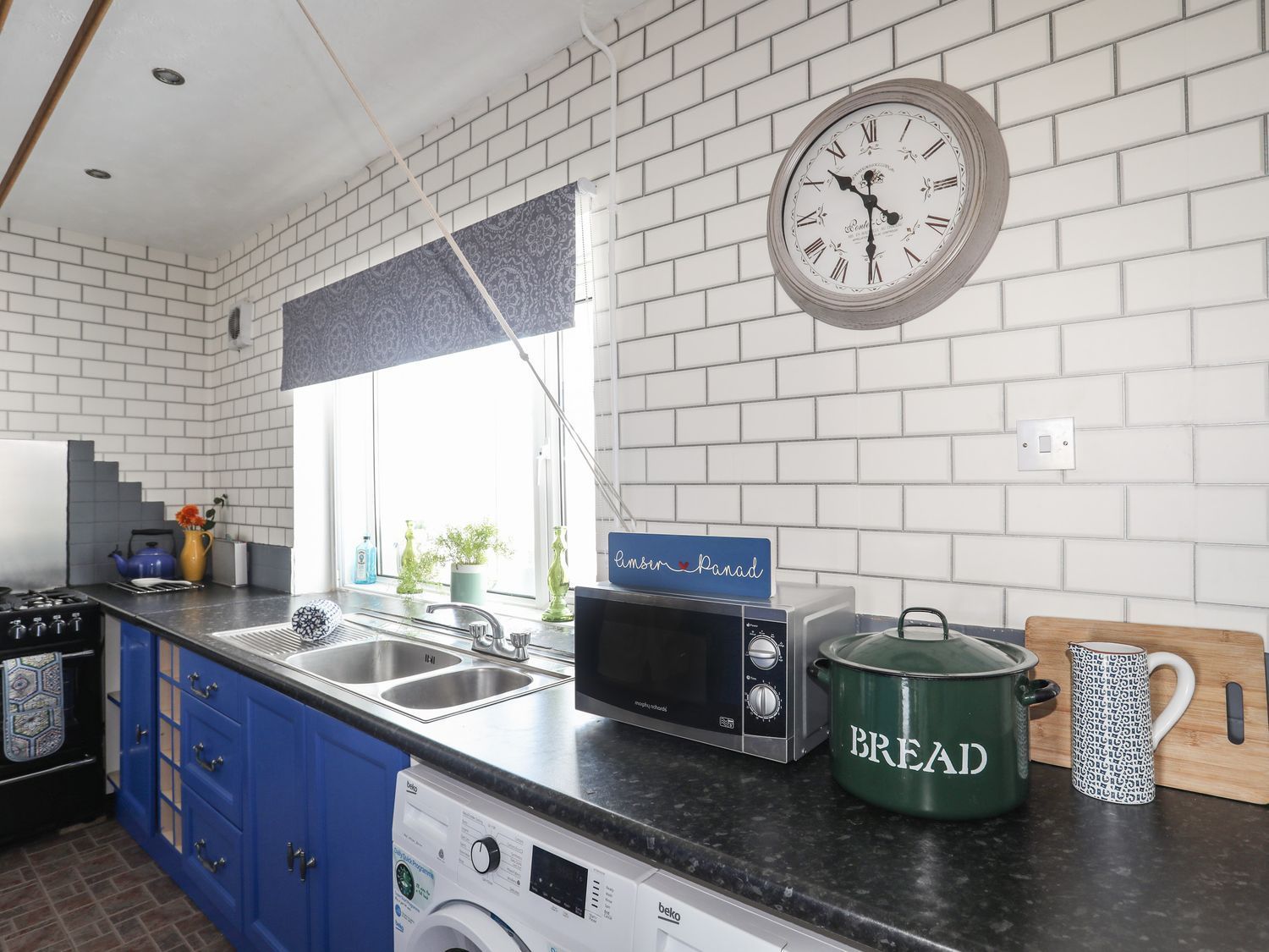 Blue kitchen with sink, microwave, bread jar, and clock on white brick wall